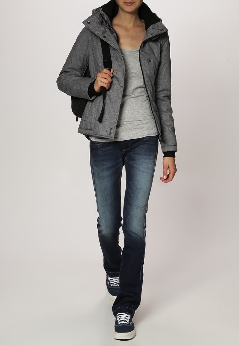 Femme marchant portant une veste grise, un t-shirt gris, un jean bleu, des baskets bleu marine, et portant un sac à dos noir sur une épaule.