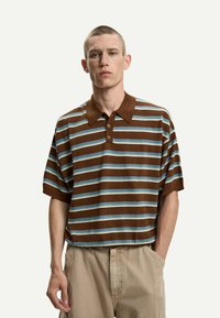 Polo a righe marroni e blu con colletto, maniche corte, tre bottoni sulla pattina e una vestibilità comoda. Indossato con pantaloni beige.
