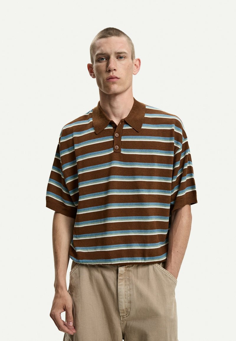 Polo a righe marroni e blu con colletto, maniche corte, tre bottoni sulla pattina e una vestibilità comoda. Indossato con pantaloni beige.