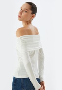 Witte off-shoulder top met geplooide stofaccenten langs de halslijn en lange, aansluitende mouwen, gecombineerd met blauwe denim jeans.