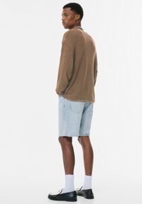 Braunfarbener, langärmeliger, strukturierter Oberteil, hellblaue Denim-Shorts, weiße Socken und schwarze Slipper. Das Model steht mit dem Rücken zu einem weißen Hintergrund.
