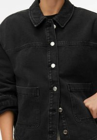 Crna traper jakna s ovratnikom, zatvaranjem na gumbe sprijeda, dva džepa na prsima i podvrnutim rukavima. Glatka tekstura s minimalnim detaljima šivanja.