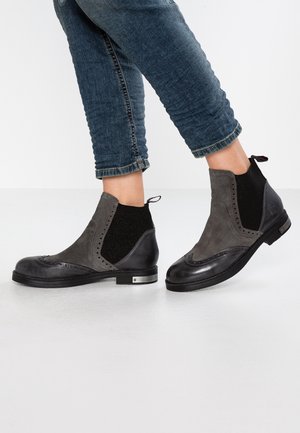 Person iført mørkegrå og sorte læder-ankelstøvler med brogue-detaljer og mørkeblå opbukne jeans, stående på hvidt gulv.