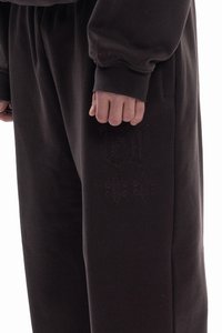 Sötétbarna melegítőnadrág elasztikus derékpánttal, oldalzsebekkel és tónusban illeszkedő hímzett díszítéssel, amely stilizált betűket és fleur-de-lis motívumot ábrázol.