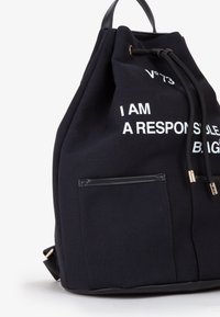 Zwarte canvas rugzak met trekkoordsluiting en contrasterende witte tekst. Heeft een voorzak met leren rand en gouden hardware.