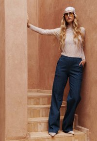 Haut à manches longues en dentelle blanche, pantalon large à rayures bleues et chaussures blanches à bout pointu. Cheveux coiffés en vagues lâches, portant des lunettes teintées de jaune.