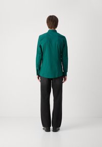 Chemise teal à manches longues avec une coupe droite, des poignets à bouton et une texture lisse, associée à un pantalon noir à jambe droite. Design simple sans motifs.