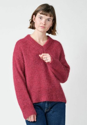 Jeune femme aux cheveux courts et bruns porte un pull à col en V rose foncé tout doux et un jean bleu, posant devant un fond clair uni.