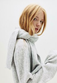 Grijze sjaal gemaakt van zacht, gestructureerd materiaal, draped om de nek. De achtergrond is licht, wat de kleur en textuur van het product benadrukt.