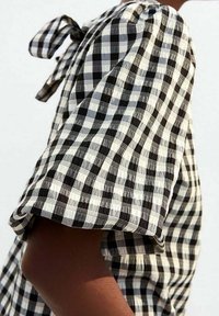 Camicia a scacchi bianca e nera con vestibilità ampia, maniche corte e dettaglio cravatta sul retro. Il tessuto appare strutturato.