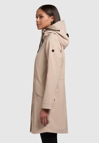 Beige vattentät jacka med huva, med en ribbad krage, framfickor och svarta detaljer. Rakt snitt, tillverkad av slät tyg.