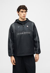 Νεαρός άνδρας που φοράει μαύρο μπουφάν Nike Shox με κουκούλα, φερμουάρ ανοιχτό μέχρι το μέσο του στήθους, και μαύρο παντελόνι, στέκεται μπροστά σε απλό φόντο.