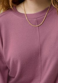 Różowa bluza z prążkowanym dekoltem i widocznymi przeszyciami. Na szyi noszony jest złoty skręcany łańcuch, dodający kontrastu.