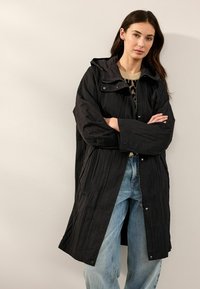 Manteau long noir, léger, avec une finition texturée, une fermeture éclair à l'avant et une capuche. Le modèle le porte sur un pull en maille et un jean.