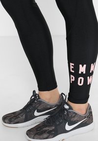 Mustat legginsit, joissa lukee "EMPOWER" vaaleanpunaisella, yhdistettynä harmaisiin ja mustiin kuvioituihin urheilukenkiin, vaalealla taustalla.