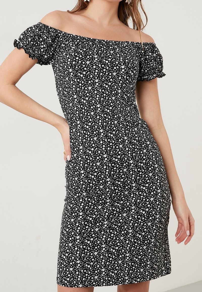 Femme portant une robe noire à épaules dénudées avec un imprimé floral blanc et des manches courtes bouffantes, posant avec une main sur la hanche.