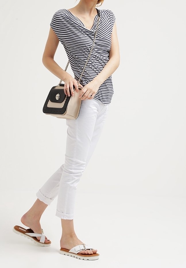 Femme portant un pantalon blanc retroussé et un haut rayé noir et blanc, chaussée de sandales blanches et tenant un sac bandoulière beige et noir à bandoulière chaîne.