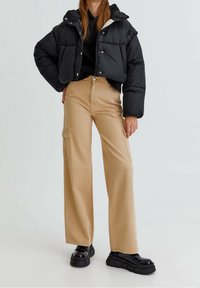 Černá péřová bunda s kapucí, khaki cargo kalhoty s bočními kapsami a masivní černé platformové boty. Hladké textury, volný střih.