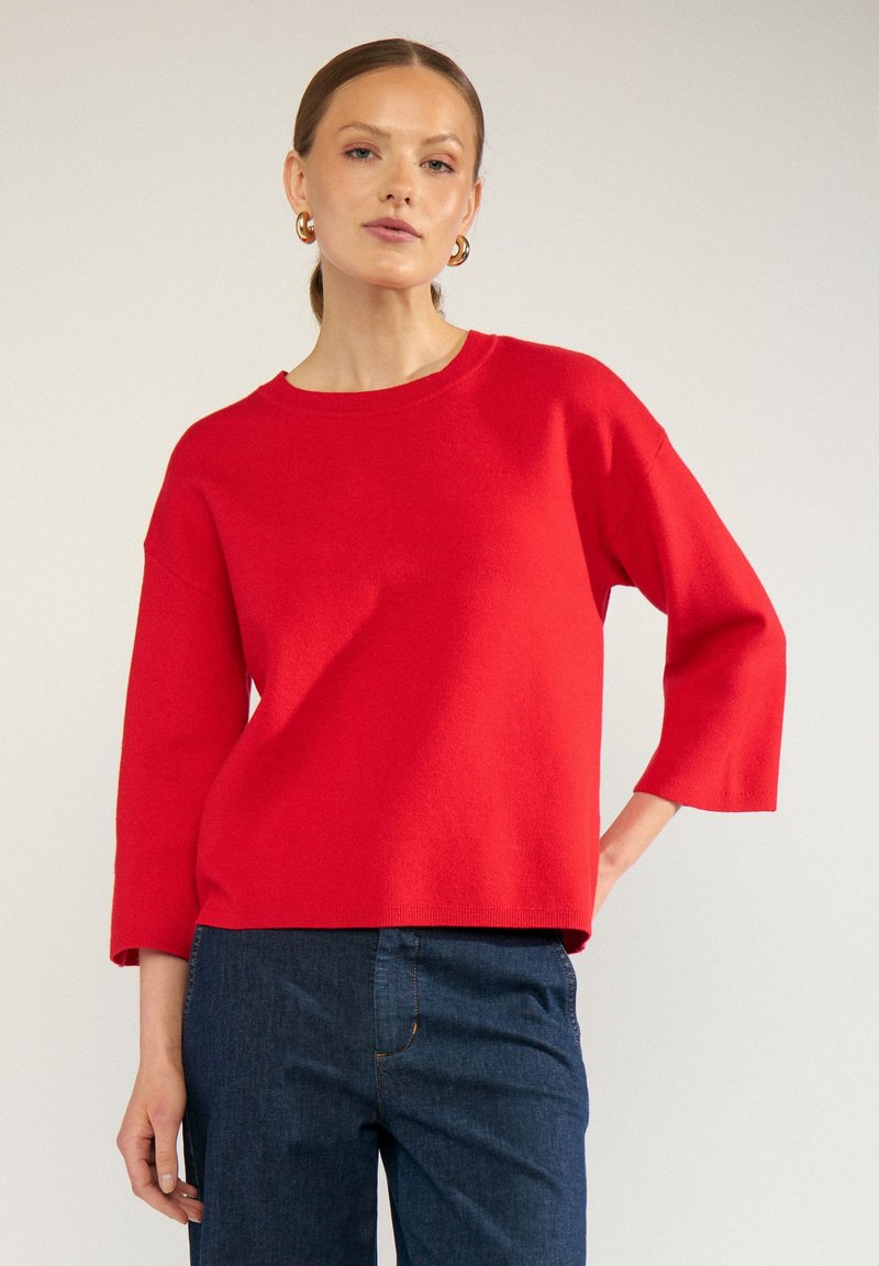 Femme portant des boucles d'oreilles créoles dorées, un pull rouge à manches longues et un jean bleu foncé, debout avec une main sur la hanche sur un fond uni.