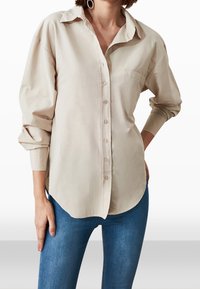 Donna che indossa una camicia beige a maniche lunghe con bottoni, tasca sul petto e jeans blu, con una mano sui fianchi e orecchini a cerchio visibili.