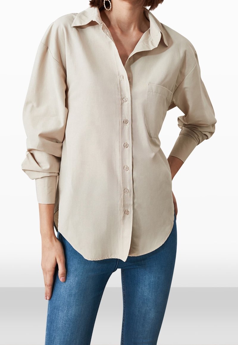 Donna che indossa una camicia beige a maniche lunghe con bottoni, tasca sul petto e jeans blu, con una mano sui fianchi e orecchini a cerchio visibili.