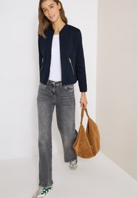 Femme portant une veste bleu marine, un haut blanc, un jean gris, des baskets vertes et blanches, tenant un grand sac à main en suède marron, debout devant un mur bicolore.
