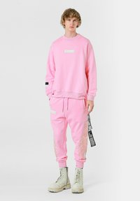 Conjunto de sudadera y pantalones de chándal en mezcla de algodón de color rosa, con una etiqueta en el pecho y detalles de bordado en los pantalones. Combinado con botas blancas.