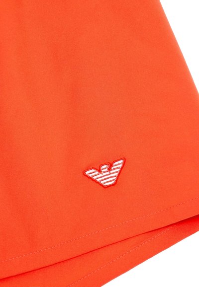 Pantalones cortos de tela naranja con un pequeño logo bordado en rojo y blanco. La textura parece suave con detalles de costura limpios.