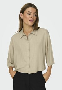 Beige kortærmet skjorte med knapper i et let, struktureret stof. Har klassisk krave og en løs pasform, båret med sorte bukser.