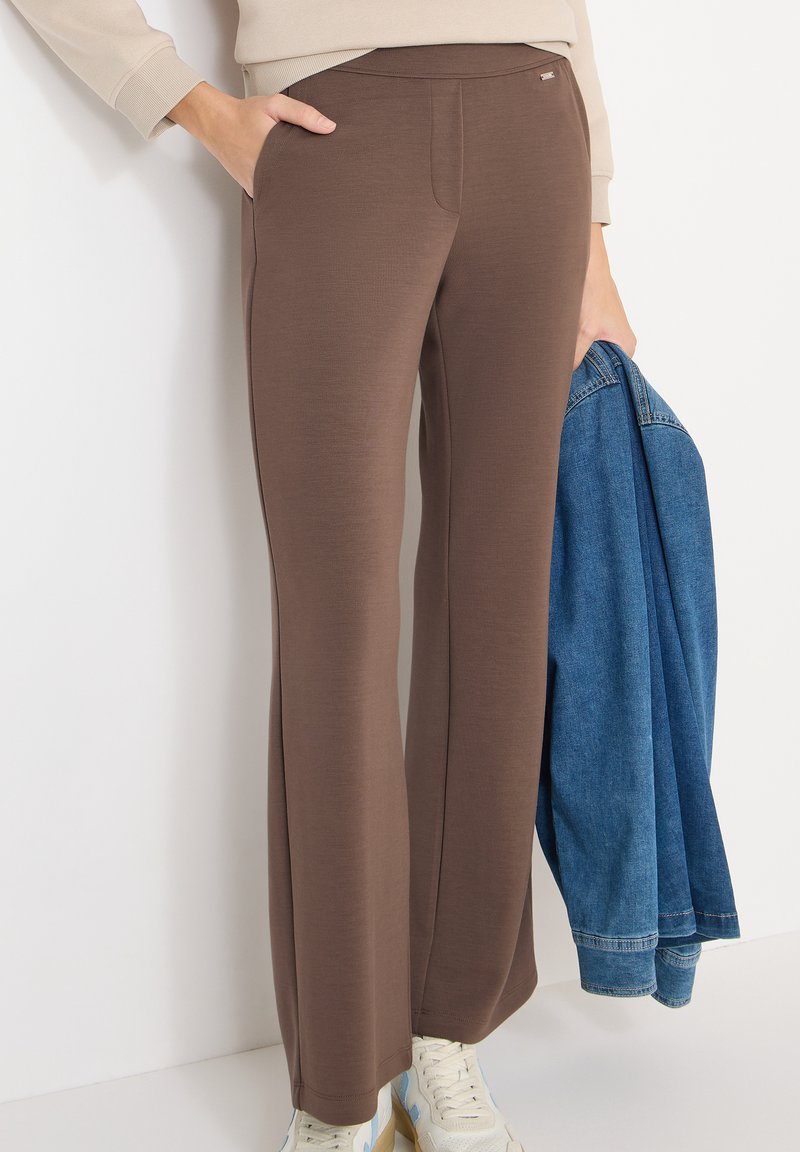 Personne portant un pantalon marron ajusté et des baskets blanches, tenant une veste en denim bleue dans une main et reposant l'autre main dans une poche.