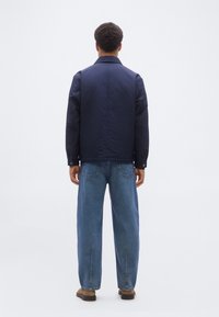 Άνδρας που στέκεται με την πλάτη γυρισμένη φορώντας μπλε ναυτικό μπουφάν, μπλε τζιν με πίσω τσέπες και καφέ παπούτσια, μπροστά σε ένα απλό λευκό φόντο.