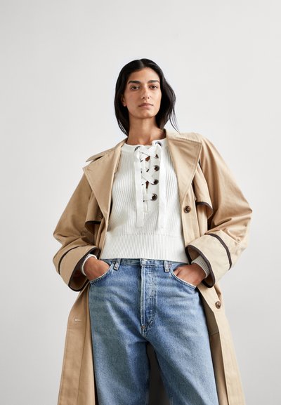 Beige trenchcoat over een wit gebreid top met een vetersluiting aan de voorkant, gecombineerd met lichtblauwe denim jeans. Glad materiaal, casual pasvorm en gedefinieerde mouwen.