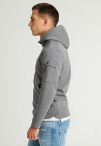 Felpa grigia con cappuccio e coulisse, caratterizzata da un orlo spaccato e tasche laterali con zip. Texture morbida e design aderente, indossata sopra jeans azzurri chiari.