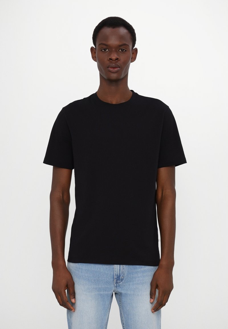 T-shirt en coton noir, à manches courtes, col rond, coupe décontractée. Porté avec un jean bleu clair, présenté sur un fond blanc uni.