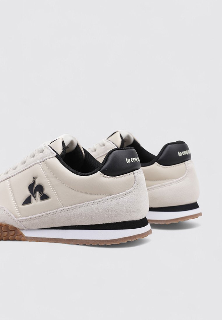 Zapatillas de cuero y ante en color beige con acentos en negro, que presentan una suela de goma texturizada, una media suela blanca y un logo en el lateral y el talón.