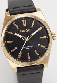 Black wristwatch with gold-toned case and markers, white hour and minute hands, and leather strap. Features water resistance up to 5 bar.