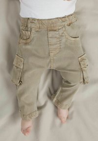 Beežid sametpüksid elastse vöökohaga, millel on kaks küljetaskut ja õmblustega detailid, asetatud helele neutraalsele taustale.