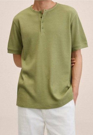 Mand iført grøn kortærmet henley-shirt med tre knapper, kombineret med hvide bukser, hånd i lommen, neutralt baggrund.
