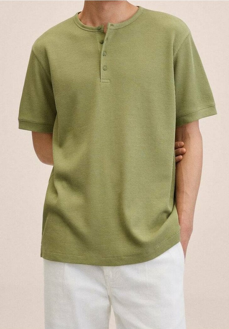 Homme portant un t-shirt henley vert à manches courtes avec trois boutons, associé à un pantalon blanc, main dans la poche, fond neutre.