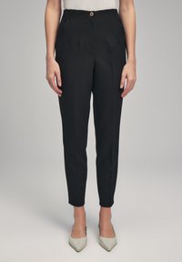 Zwarte op maat gemaakte broek met voorplooien, zakken en een gouden knoopsluiting; slim fit, enkel-lange ontwerp gecombineerd met lichtgekleurde hakken.
