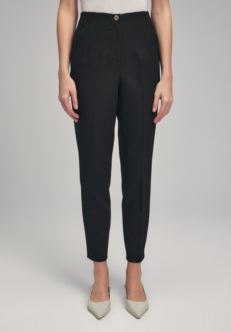 Černé přizpůsobené kalhoty s předními sklady, kapsami a zlatým knoflíkem; slim fit, po kotníky, doplněné světlými podpatky.