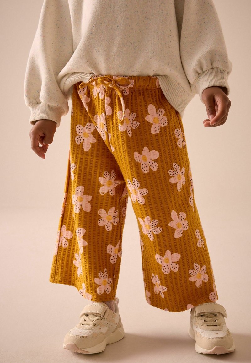 Enfant portant un pantalon large à fleurs jaune moutarde, un pull crème oversize et des baskets blanc cassé, debout devant un fond uni.