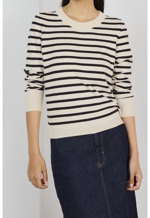 Femme portant un pull à manches longues rayé horizontalement crème et noir avec une jupe en jean bleu foncé.