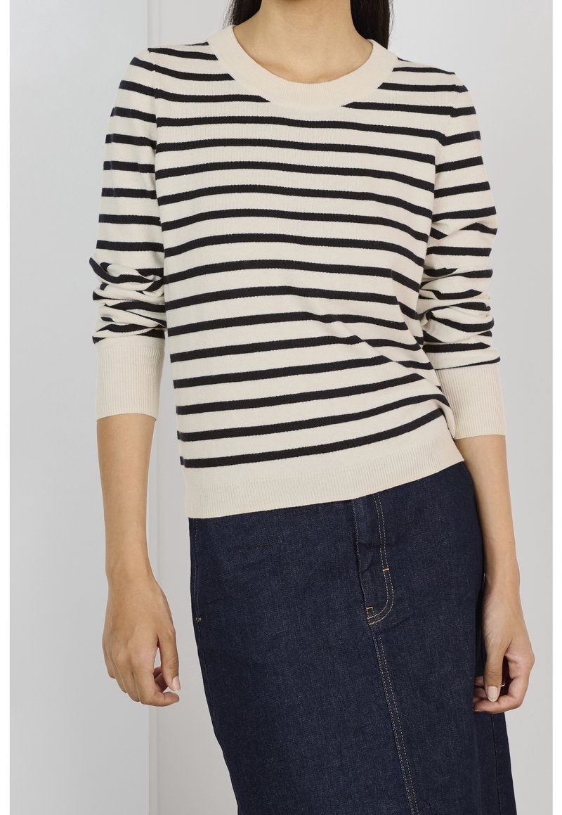 Femme portant un pull à manches longues rayé horizontalement crème et noir avec une jupe en jean bleu foncé.