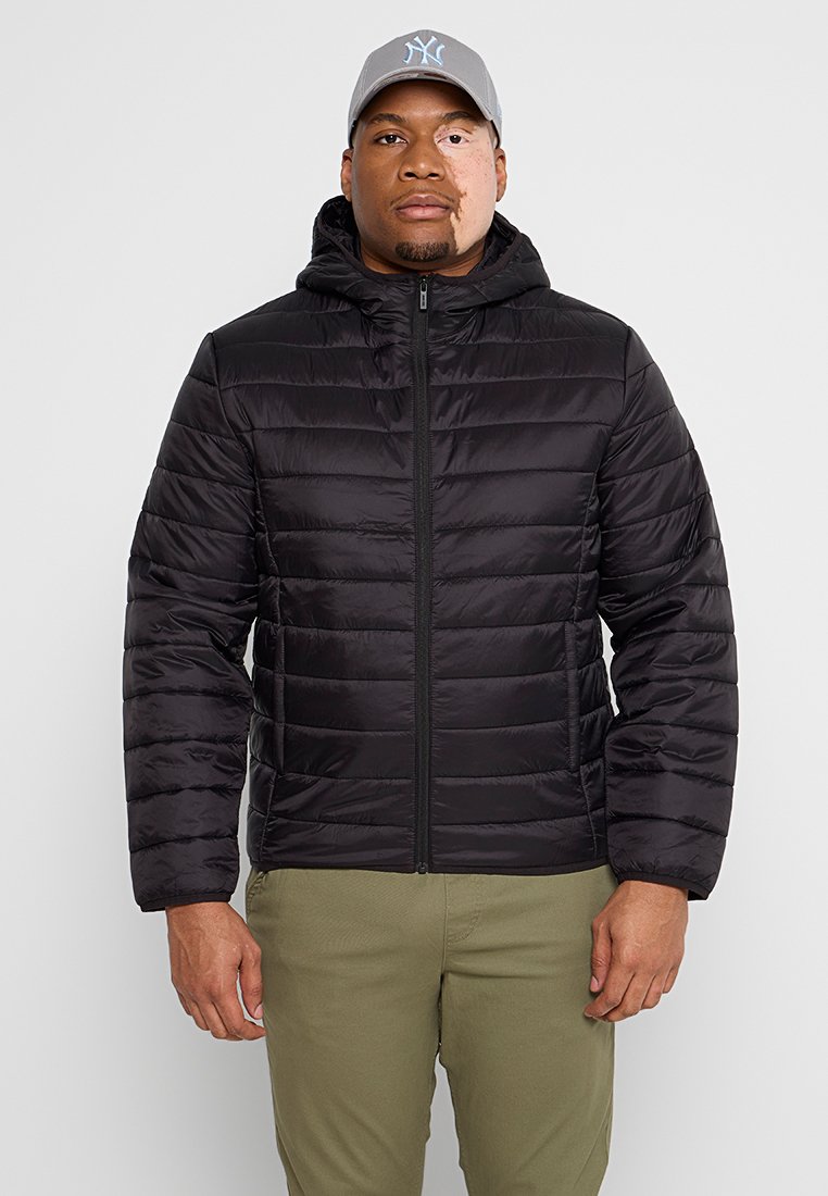 Giacca a vento nera con chiusura a zip, design trapuntato e cappuccio. Indossata sopra pantaloni verde oliva, abbinata a un cappellino grigio con logo.