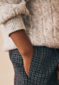 Grijze kabelgebreide trui met omgeslagen mouwen, gecombineerd met donkere geruite broek. Hand in de zak gestoken, met zichtbaar gladde huidtextuur.