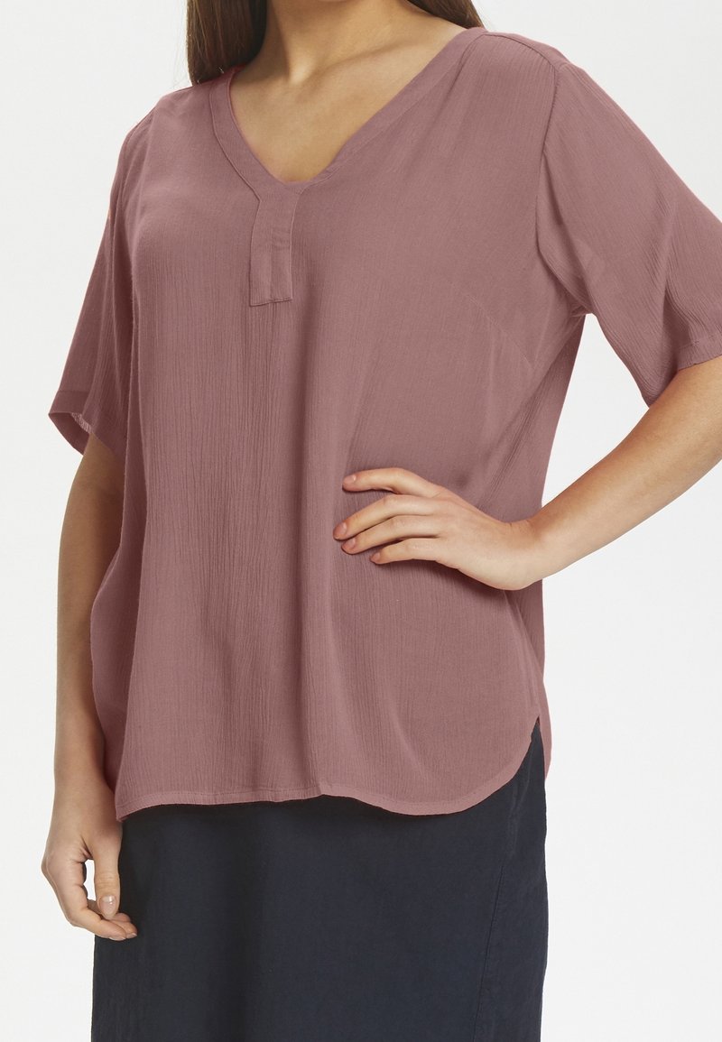 Kortærmet, blushing pink bluse med V-udskæring og en struktureret overflade. Har en lille frontplacket og afrundet kant, parret med en mørk nederdel.