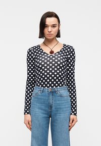 Γυναίκα με κοντά σκουρόχρωμα μαλλιά, που φοράει μπλε μπλούζα με μακριά μανίκια και λευκές πουά, τζιν ψηλόμεσο μπλε και κολιέ με μενταγιόν σε σχήμα τριαντάφυλλου.