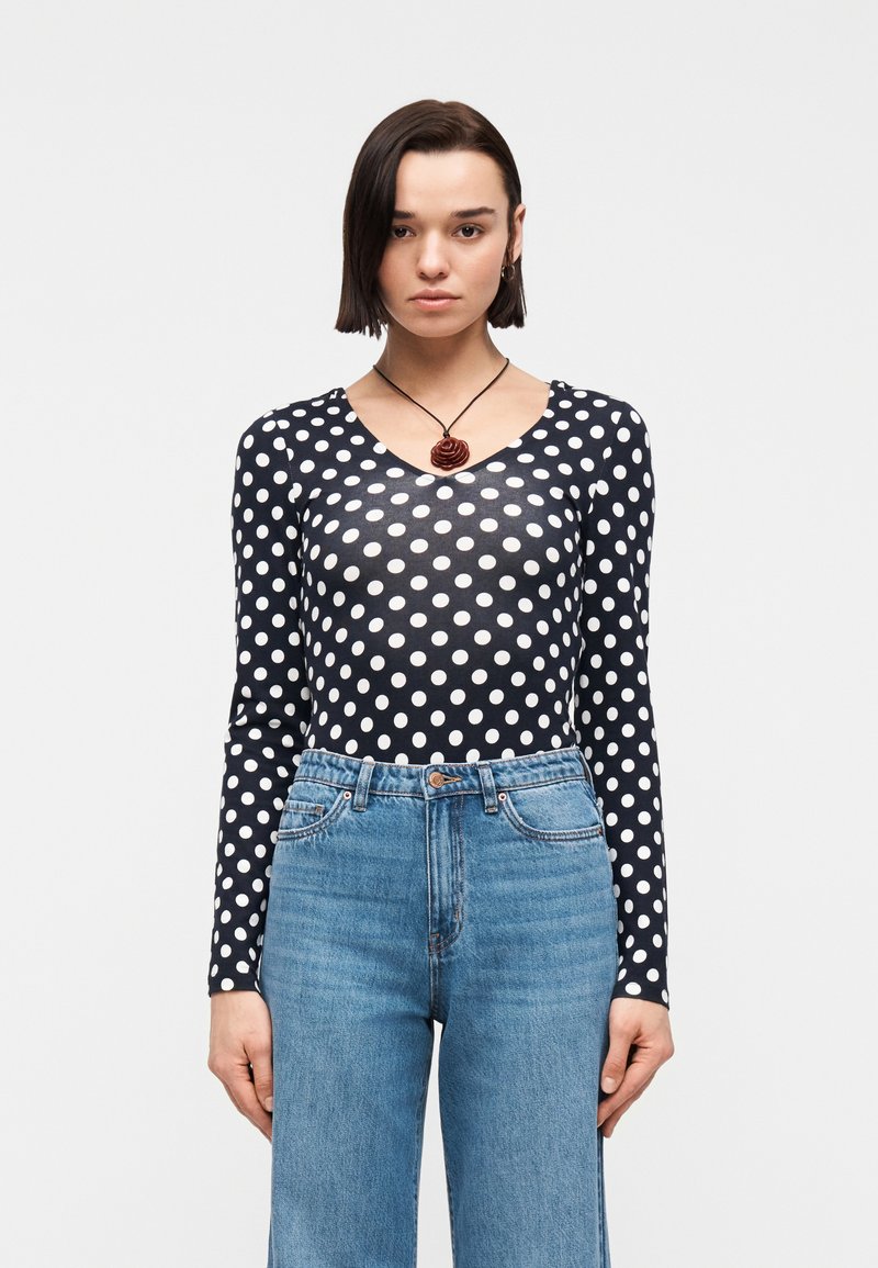 Γυναίκα με κοντά σκουρόχρωμα μαλλιά, που φοράει μπλε μπλούζα με μακριά μανίκια και λευκές πουά, τζιν ψηλόμεσο μπλε και κολιέ με μενταγιόν σε σχήμα τριαντάφυλλου.