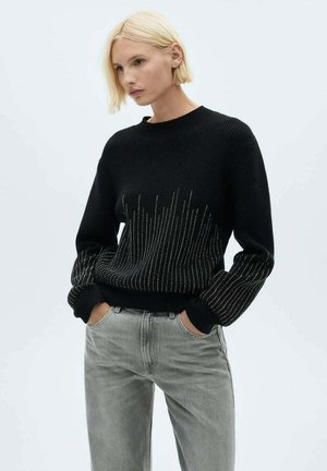 Schwarzer Strickpullover mit gerippter Textur und metallischen vertikalen Akzentlinien. Kombiniert mit hellgrauen Jeans.